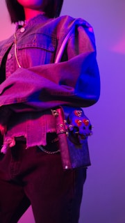 A person wearing a denim jacket and black pants, holding a uniquely designed bag with red embellishments. The lighting casts vibrant shades of purple and pink, creating a striking contrast.