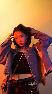 A person is standing against a warm, gradient-lit background, wearing a denim jacket over a black top. Their hands are placed on their head, and they have an intense, contemplative gaze.
