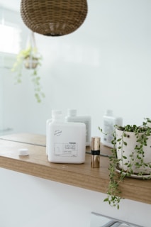 A minimalist bathroom shelf holds a white bottle labeled 'TONIK', a small gold cylindrical container, and a potted plant with trailing vines. Above, there is a woven basket-style light fixture. The background is mostly white, creating a clean and airy atmosphere.