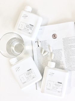 Three white bottles labeled 'TONIK' are arranged on a white surface beside a clear glass of water. They are placed on an open magazine showing black and white text, along with a few scattered capsules. The scene is minimalistic and clean, with an emphasis on wellness or lifestyle products.