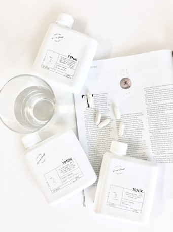 Three white bottles labeled 'TONIK' are arranged on a white surface beside a clear glass of water. They are placed on an open magazine showing black and white text, along with a few scattered capsules. The scene is minimalistic and clean, with an emphasis on wellness or lifestyle products.