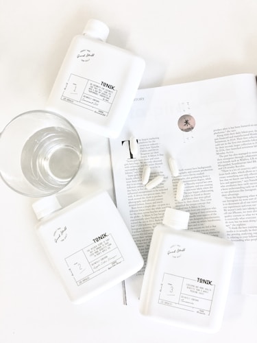 Three white bottles labeled 'TONIK' are arranged on a white surface beside a clear glass of water. They are placed on an open magazine showing black and white text, along with a few scattered capsules. The scene is minimalistic and clean, with an emphasis on wellness or lifestyle products.
