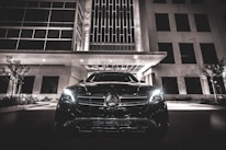 A luxury car with headlights on is parked in front of a modern building at night. The building features geometric windows and a canopy over the entrance. The scene is illuminated by artificial lights, creating a sleek and sophisticated atmosphere.
