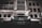 A luxury car with headlights on is parked in front of a modern building at night. The building features geometric windows and a canopy over the entrance. The scene is illuminated by artificial lights, creating a sleek and sophisticated atmosphere.