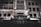 A luxury car with headlights on is parked in front of a modern building at night. The building features geometric windows and a canopy over the entrance. The scene is illuminated by artificial lights, creating a sleek and sophisticated atmosphere.