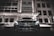 A luxury car with headlights on is parked in front of a modern building at night. The building features geometric windows and a canopy over the entrance. The scene is illuminated by artificial lights, creating a sleek and sophisticated atmosphere.