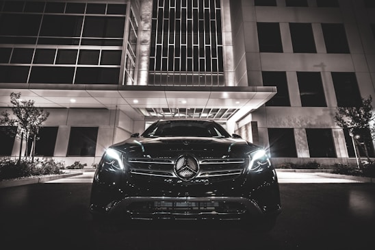 A luxury car with headlights on is parked in front of a modern building at night. The building features geometric windows and a canopy over the entrance. The scene is illuminated by artificial lights, creating a sleek and sophisticated atmosphere.