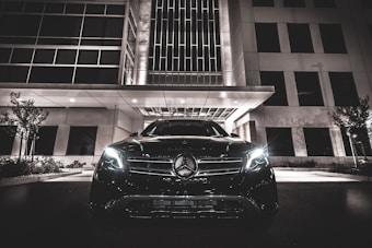 A luxury car with headlights on is parked in front of a modern building at night. The building features geometric windows and a canopy over the entrance. The scene is illuminated by artificial lights, creating a sleek and sophisticated atmosphere.