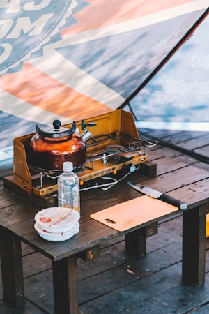 A wooden table holds a portable gas stove with a kettle on it, a plastic water bottle, a knife, a cutting board, and two stacked food containers. The background features a slightly blurred tent with a graphic design.
