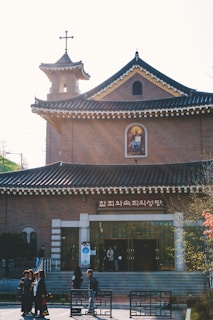 A brick church building features traditional architecture with a curved, tiled roof and a small tower topped by a cross. A religious icon is displayed prominently above the entrance. Several people are gathered at the entrance and walking nearby. The sunny sky casts a bright light over the scene.