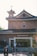 A brick church building features traditional architecture with a curved, tiled roof and a small tower topped by a cross. A religious icon is displayed prominently above the entrance. Several people are gathered at the entrance and walking nearby. The sunny sky casts a bright light over the scene.