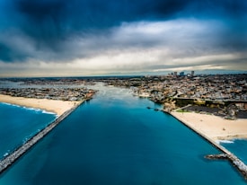 Aerial view of a coastal city, featuring a wide body of calm, blue water leading from the ocean into the city. Sand beaches line both sides of the water, and numerous buildings populate the landscape, tapering off into the distance. The sky is cloudy, with a mix of dark and light clouds adding a dramatic effect.