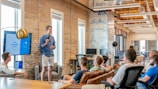 A group of people are seated in a modern office setting, attentively listening to a man standing and speaking in front of them. The speaker is holding a device, possibly a remote or microphone, and there is a presentation displayed on a screen behind him. The room is well-lit with natural light coming through large windows, and the decor includes exposed brick walls and modern furniture.