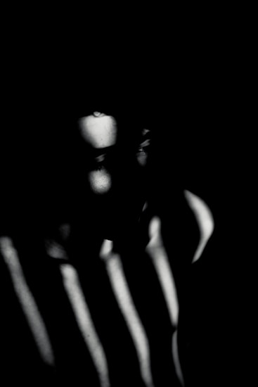 A dark portrait with intense shadows and light creating a dramatic contrast over a human face. The play of light and shadow obscures the features, giving an enigmatic and mysterious appearance.