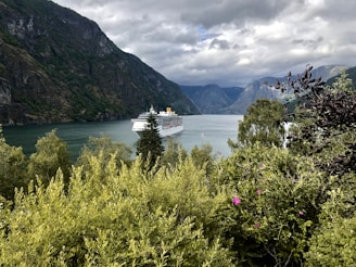 A cruise ship sails through a serene fjord surrounded by towering mountains and lush greenery. The sky is overcast with clouds, adding a dramatic contrast to the landscape. Bright green foliage in the foreground frames the view, with pink flowers adding a splash of color.