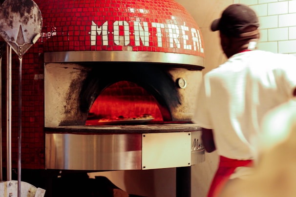 A pizza is being cooked inside a traditional, dome-shaped brick oven with red tiles and the word 'Montreal' on it. A person wearing a white shirt and a red apron is in the foreground, possibly a chef or baker, attending to the oven. There is also a metal pizza peel leaning against the oven.