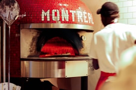 A pizza is being cooked inside a traditional, dome-shaped brick oven with red tiles and the word 'Montreal' on it. A person wearing a white shirt and a red apron is in the foreground, possibly a chef or baker, attending to the oven. There is also a metal pizza peel leaning against the oven.