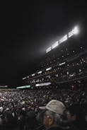 A large stadium filled with a crowd of spectators, illuminated by bright stadium lights. The seating areas are packed with attendees, many of whom are wearing caps and jackets. The scoreboard is visible, displaying scores, and various advertisements are lit up along the stands.