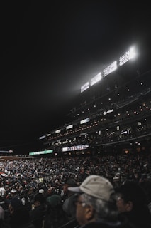 A large stadium filled with a crowd of spectators, illuminated by bright stadium lights. The seating areas are packed with attendees, many of whom are wearing caps and jackets. The scoreboard is visible, displaying scores, and various advertisements are lit up along the stands.