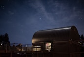 A modern observatory with a curved metallic roof is illuminated under a starry night sky. Soft lighting emits from inside the building, and shadows of nearby trees and fencing create a serene atmosphere.