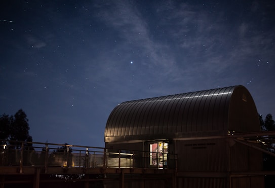 A modern observatory with a curved metallic roof is illuminated under a starry night sky. Soft lighting emits from inside the building, and shadows of nearby trees and fencing create a serene atmosphere.
