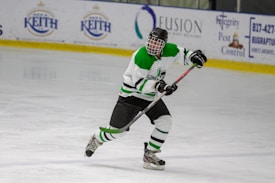 A hockey player in green and white gear is actively skating on an ice rink, gripping a hockey stick. Advertisements are visible along the edge of the arena.