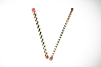 Two makeup brushes are placed on a white background, crossing each other to form a V shape. Both brushes have metallic handles with soft brown bristles, and one of the brushes has a slight pink tint on the bristles.