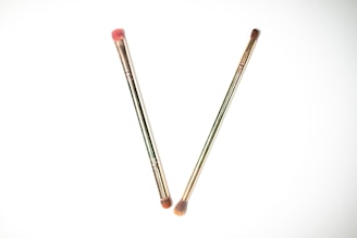 Two makeup brushes are placed on a white background, crossing each other to form a V shape. Both brushes have metallic handles with soft brown bristles, and one of the brushes has a slight pink tint on the bristles.