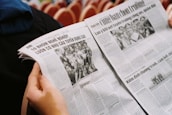 A person is holding an open newspaper with articles and black-and-white photographs. The text appears to be in Vietnamese, and the layout includes headlines, columns, and images of people.