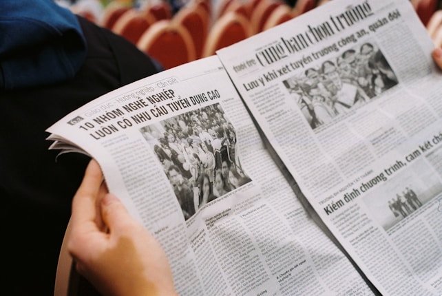 A person is holding an open newspaper with articles and black-and-white photographs. The text appears to be in Vietnamese, and the layout includes headlines, columns, and images of people.