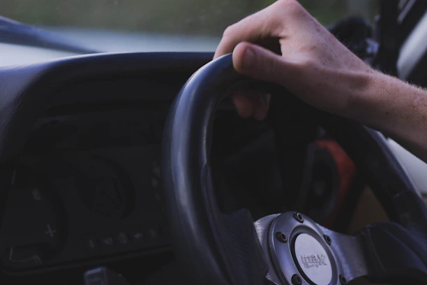 Close-up of a driver's hand holding a steering wheel with a city street in the background.