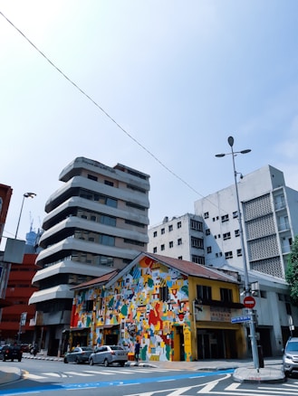 A vibrant street scene with a colorful mural covering the facade of a small building, featuring abstract and geometric patterns. Surrounding it are modern, multi-story buildings with unique architectural designs. Several cars are parked along the street, and there are road signs visible.