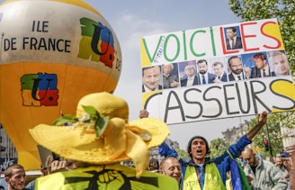A group of people are gathered at a protest holding signs and a large balloon with 'ILE DE FRANCE' written on it. One of the signs has pictures of several individuals and the text 'VOICI LES CASSEURS'. The crowd appears engaged in expressing dissent or discontent.