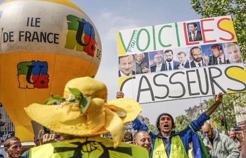 A group of people are gathered at a protest holding signs and a large balloon with 'ILE DE FRANCE' written on it. One of the signs has pictures of several individuals and the text 'VOICI LES CASSEURS'. The crowd appears engaged in expressing dissent or discontent.