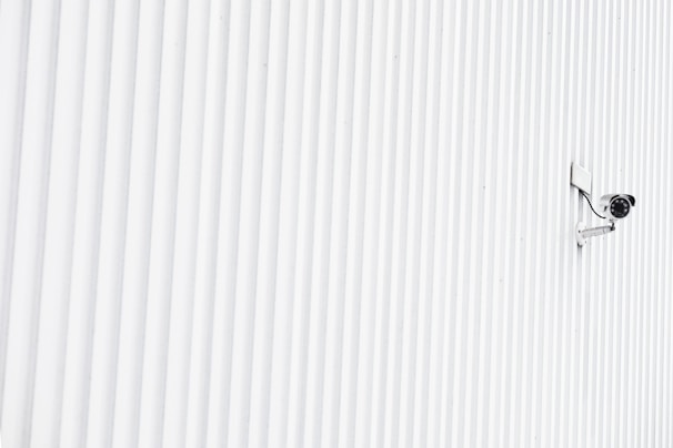 A solitary security camera is mounted on a white, ribbed wall that spans the entire image, creating a minimalist and modern aesthetic.