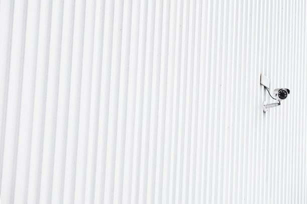 A solitary security camera is mounted on a white, ribbed wall that spans the entire image, creating a minimalist and modern aesthetic.