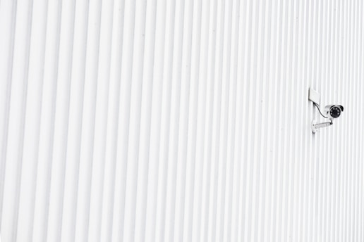 A solitary security camera is mounted on a white, ribbed wall that spans the entire image, creating a minimalist and modern aesthetic.