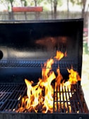 A barbecue grill with bright, orange flames rising from the grill grates. The background is slightly blurred with hints of greenery and a wooden fence, indicating an outdoor setting.