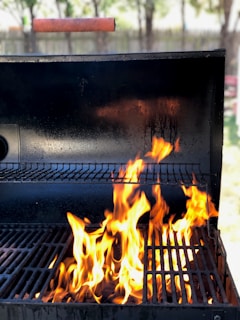 A barbecue grill with bright, orange flames rising from the grill grates. The background is slightly blurred with hints of greenery and a wooden fence, indicating an outdoor setting.
