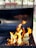 A barbecue grill with bright, orange flames rising from the grill grates. The background is slightly blurred with hints of greenery and a wooden fence, indicating an outdoor setting.