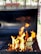 A barbecue grill with bright, orange flames rising from the grill grates. The background is slightly blurred with hints of greenery and a wooden fence, indicating an outdoor setting.