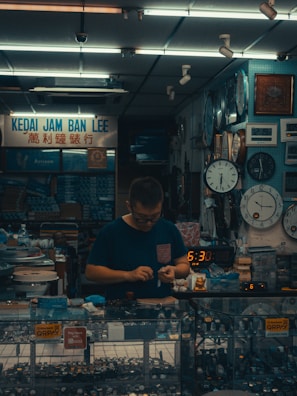 A small watch shop with various clocks displayed on the wall. A person is standing behind a glass counter filled with wristwatches, focusing on handling a small object. The setting suggests a cozy, somewhat cluttered workspace filled with timepieces.