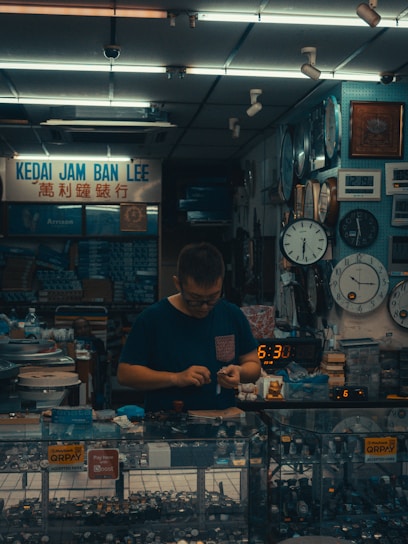 A small watch shop with various clocks displayed on the wall. A person is standing behind a glass counter filled with wristwatches, focusing on handling a small object. The setting suggests a cozy, somewhat cluttered workspace filled with timepieces.