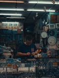 A small watch shop with various clocks displayed on the wall. A person is standing behind a glass counter filled with wristwatches, focusing on handling a small object. The setting suggests a cozy, somewhat cluttered workspace filled with timepieces.