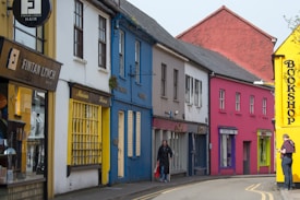 A quaint street scene features several colorful buildings with a variety of vibrant hues. The storefronts include a hair salon, a yellow-painted building labeled Marian House, and a bookshop with a bright yellow facade. The street is slightly curved, and a few pedestrians are visible, including one with a walking stick and another using a mobile phone.