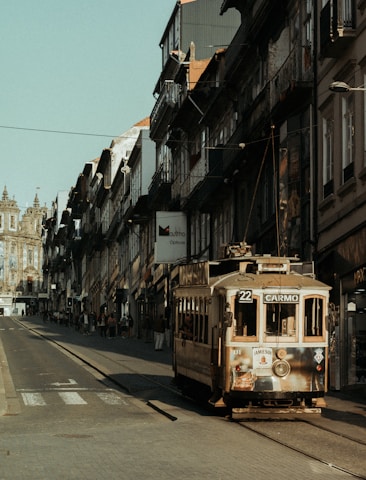 A vintage tram labeled '22 Carmo' travels along a narrow street lined with historic buildings. The architecture features intricate facades and balconies, with some people walking on the cobblestone sidewalk and a distant view of a large, ornate building with spires.
