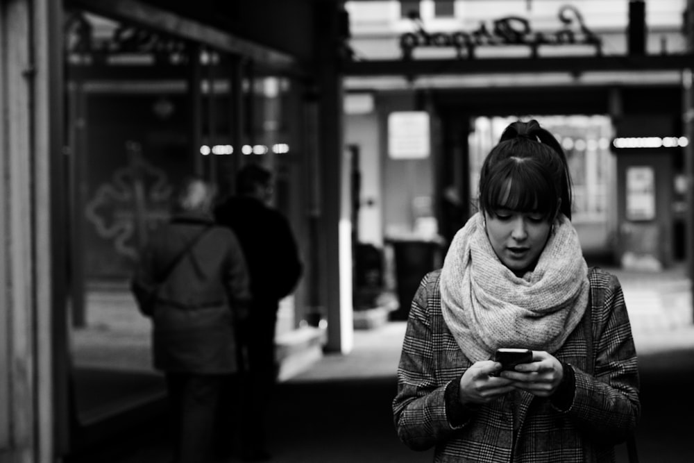 grayscale photography of woman walking near street while using smartphone