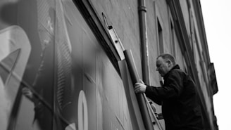 A man is standing on a ladder next to a building, holding a rolled-up poster or sheet. The building facade is partially in view, with a pipe running parallel to the ladder. The scene is captured in black and white, giving it a classic and timeless feel.