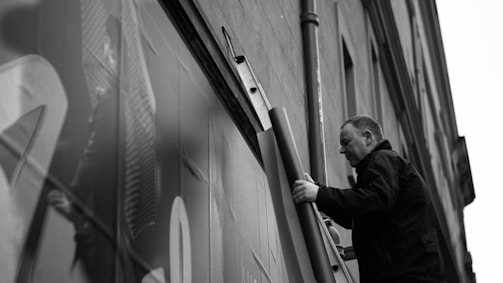 A man is standing on a ladder next to a building, holding a rolled-up poster or sheet. The building facade is partially in view, with a pipe running parallel to the ladder. The scene is captured in black and white, giving it a classic and timeless feel.
