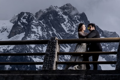 A couple, dressed in formal attire, is standing on a bridge against the backdrop of snow-covered mountains. The woman is wearing a wedding dress and is gently holding the man's cheek, while he leans in close to her, both appearing happy.